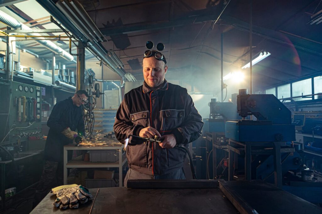 Handyman preparing his autogenous welding tool for work while his colleague can be noticed in the back working on his grinding task.